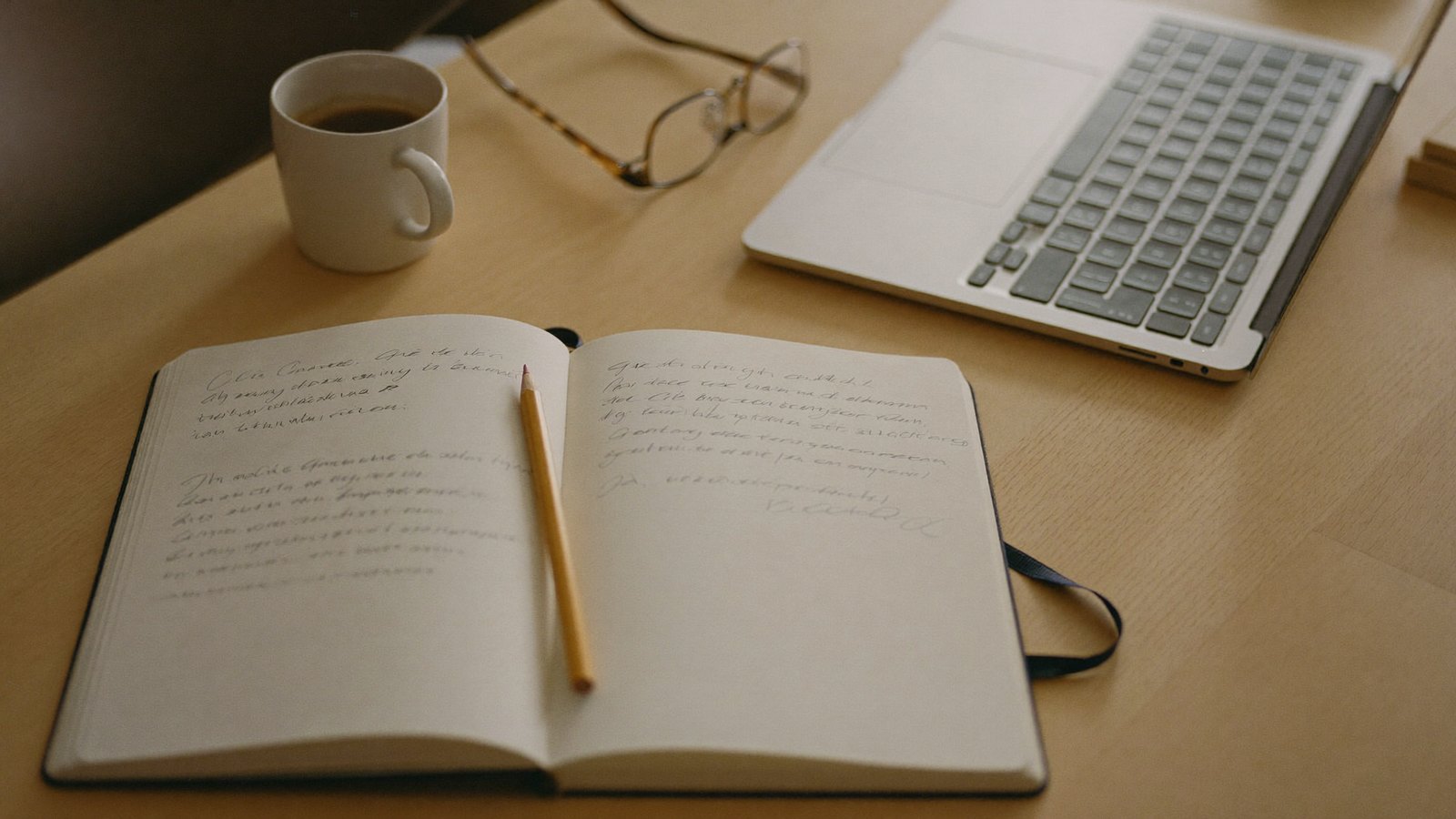 An open notebook with handwritten notes beside a laptop on a light-wood desk in soft morning light — a calm, careful, doing-the-homework still life.
